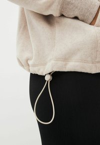 Sudadera de felpa color beige claro con puños acanalados y un cordón ajustable en el dobladillo, combinada con leggings acanalados negros.