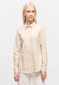 Woman with straight blonde hair wearing a beige and white gingham button-up shirt and white pants, standing against a plain white background.