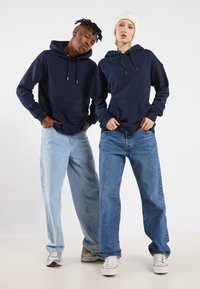 To personer har på seg marineblå hettegensere med lommer foran, kombinert med løstsittende blå jeans. Hettegenserne har snorer og en uformell design.