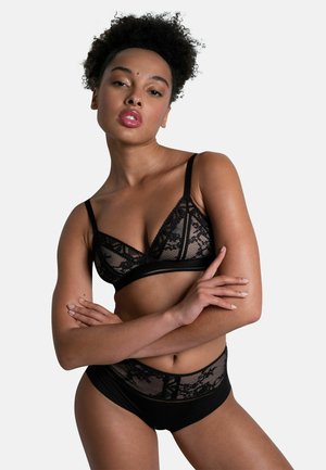 Vrouw met krullend haar die een zwarte kanten beha en bijpassende hoge taille slip draagt, staande met gekruiste armen tegen een effen achtergrond.