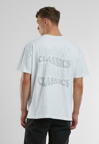 Baltas kokvilnas T-krekls ar pārspīlētu "URBAN CLASSICS" tekstu biezā, ēnu stilā uz muguras, īsām piedurknēm un brīvā stilā.