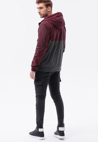 Mörkröd och grå zip-up jacka med huva, tillverkad av ett lättviktigt, glänsande tyg. Parat med figurnära svarta cargobyxor och svarta sneakers.