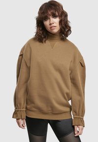 Jeune femme aux cheveux bruns bouclés portant un sweat-shirt à col roulé ample de couleur beige et un short noir brillant, sur un fond uni.