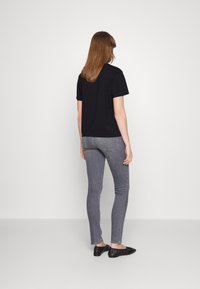 Kvinde med lige brunt hår står med ryggen til, iført en sort T-shirt med korte ærmer, grå tætsiddende jeans og sorte slip-on sko.