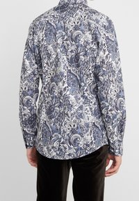 Mężczyzna noszący koszulę z długim rękawem o skomplikowanym niebiesko-białym wzorze kwiatowym paisley, w zestawieniu z czarnymi spodniami, widziany od tyłu.