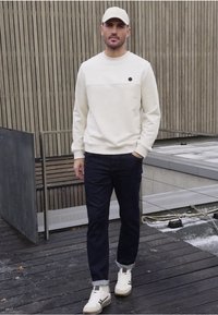 Světlé béžové mikina s horizontálním pruhem a černým akcentem, tmavě modré džíny a bílé tenisky se šedými a černými detaily, nošené s kšiltovkou.