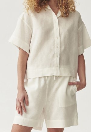 Femme portant une chemise blanche ample à manches courtes boutonnée et un short blanc assorti, main dans la poche, sur un fond uni.
