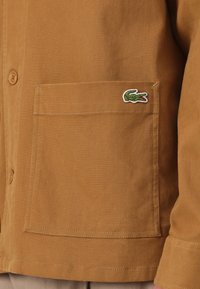 Poche de veste en velours côtelé marron avec un petit logo crocodile vert brodé près du coin supérieur droit, à côté de boutons couleur fauve.