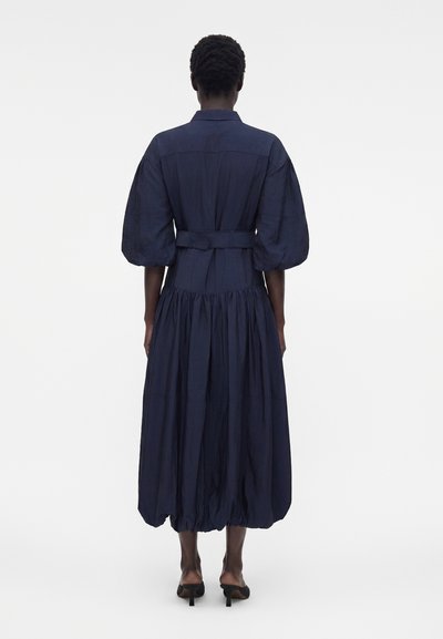 Άτομο που στέκεται με την πλάτη, φοράει ένα μίντι φόρεμα σε ναυτικό μπλε χρώμα με φουσκωτά μανίκια, εφαρμοστή μέση και ραμμένο μπαλόν φόρεμα, συνδυασμένο με μαύρες μουλάρες με τακούνι.