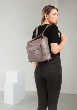 Vrouw draagt een zwart shirt en donkere spijkerbroek, met een taupekleurige leren rugzak, staand in een minimalistische grijze kamer met witte sokkelblokken.