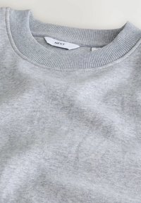 Sudadera gris con cuello redondo acanalado, fabricada en material suave. Presenta un diseño clásico y una sutil textura con una etiqueta de logo.