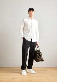 White button-up shirt, black trousers, white sneakers, and a dark green leather bag with brown accents. Casual fit with a baseball cap.