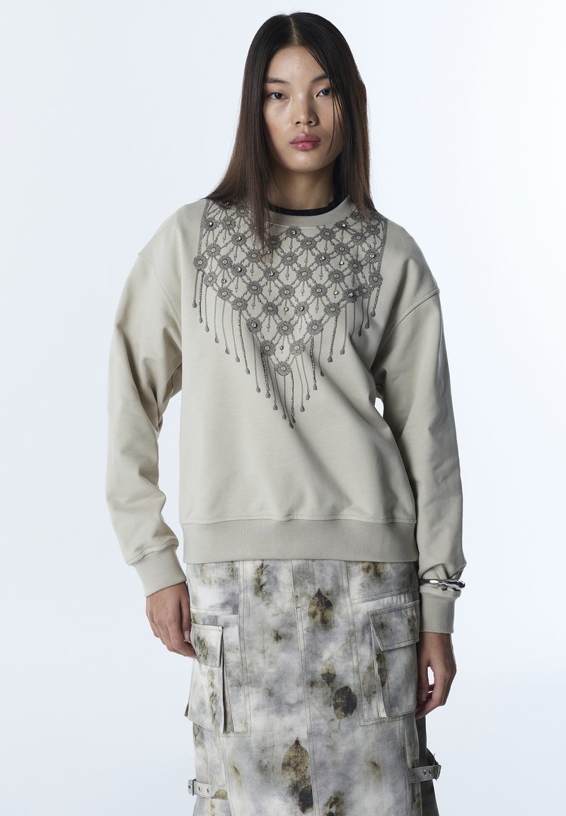 Mujer con sudadera beige con diseño detallado de encaje y cuentas en el pecho y falda cargo gris con estampado, bolsillos y correas.