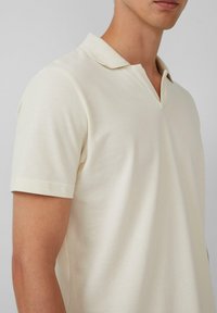 Beżowa koszulka polo z niskim dekoltem w serek, krótkimi rękawami i teksturowanym materiałem. Minimalistyczny design bez widocznych logo czy wzorów.