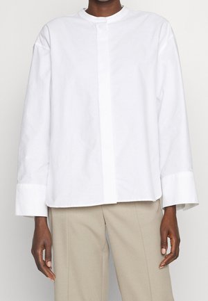 Osoba ubrana w luźną, białą koszulę z długim rękawem i szerokimi mankietami oraz beżowe, dopasowane spodnie na tle gładkiego tła.