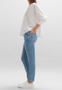 Vit oversized blus med långa ärmar, matchad med ljusblå slim-fit jeans och vita sneakers, visad från sidan mot en neutral bakgrund.