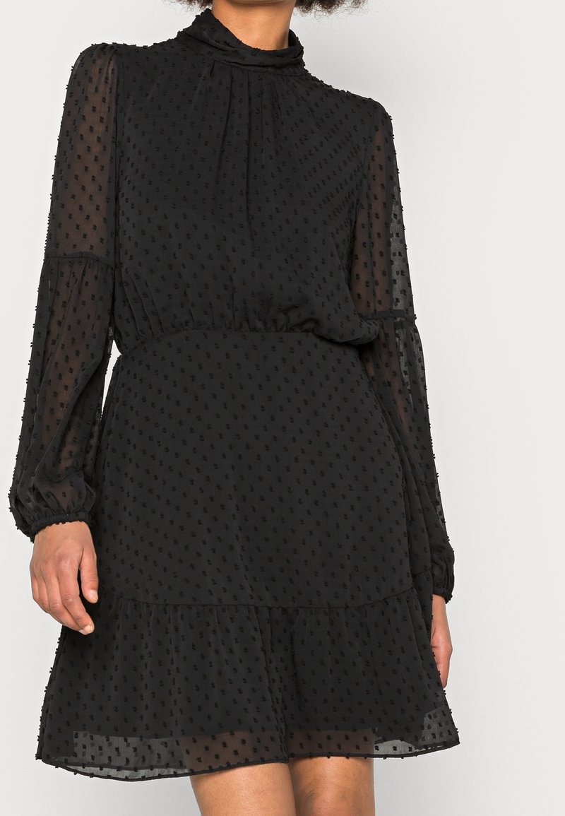 Femme portant une robe noire transparente à manches longues avec une texture à pois, un col montant, une taille élastique et un ourlet à volants, debout devant un fond uni.