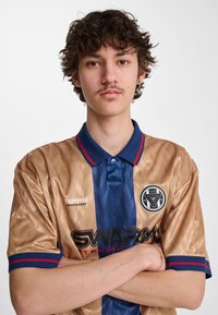 Beige og blå trøje med geometrisk mønster, med krave, korte ærmer og logoer fra "hummel" og "SWARM International".