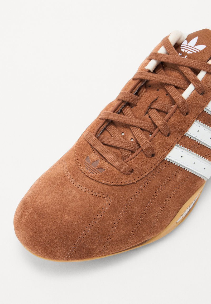 Braune Wildleder-Sportschuhe mit weißen Streifen und Markenlogo. Merkmale sind das strukturierte Obermaterial, die runde Zehenpartie und die kontrastierende Gummisohle.