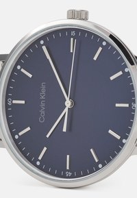 Montre Calvin Klein avec un cadran bleu marine, des index métalliques, des aiguilles argentées et un boîtier en acier inoxydable. Le texte indique "Calvin Klein" sous le marqueur des 12 heures.
