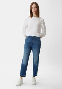 Biała koszula z długim rękawem o strukturalnej fakturze, w połączeniu z jasnoniebieskimi spodniami typu straight-leg z przetartymi miejscami i postrzępionym dołem. Białe sneakersy.