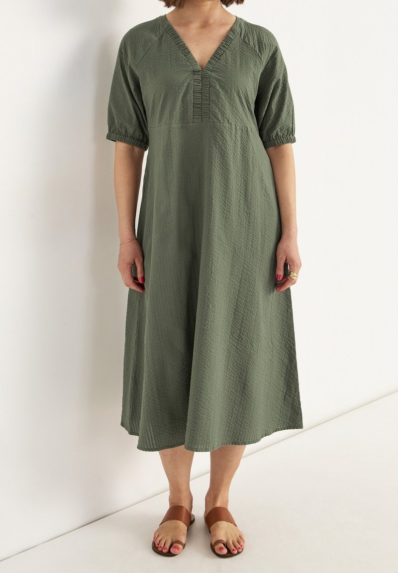 Oliwkowa sukienka midi z dekoltem w kształcie litery V, krótkimi bufkami i teksturowanym materiałem. Zestawiona z brązowymi płaskimi sandałami.