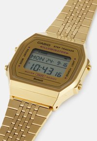 Montre de suivi de pas numérique Casio avec un boîtier rectangulaire doré, un bracelet en maillons métalliques et un écran LCD affichant l'heure et la date.