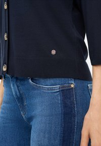 Cardigan blu navy realizzato in un tessuto a maglia fine, con colletto a girocollo, chiusura frontale con bottoni e un discreto logo vicino all'orlo. Abbinato a jeans blu.