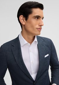 Blazer strutturato blu scuro con revers, abbinato a una camicia bianca con bottoni. Caratterizzato da un fazzoletto bianco nella tasca.