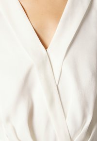 Blusa blanca envolvente con un profundo escote en V, hecha de una tela suave y ligera, con suaves pliegues y un sutil brillo.