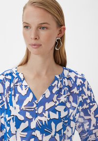 Blaue gemusterte Bluse mit abstrakten weißen und schwarzen Designs, runder Ausschnitt und weichem, leichtem Stoff. Silberne gewellte Ohrringe.