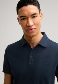 Camisa polo azul marino con cuello, mangas cortas y cierre de botón. Cuenta con un sutil logo bordado en el lado izquierdo del pecho. Textura suave.