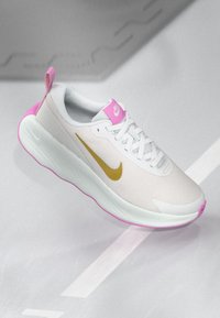 Λευκό αθλητικό παπούτσι από διχτυωτό ύφασμα με ροζ λεπτομέρειες και χρυσό σήμα Nike. Διαθέτει στρογγυλή σόλα και υφασμάτινη επιφάνεια με υφή για καλύτερη αναπνοή.