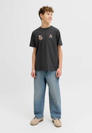 Teenagedreng iført en sort grafisk t-shirt, lyseblå jeans og beige sko, stående og kiggende til siden mod en hvid baggrund.