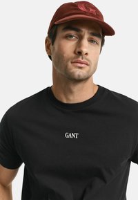 Man die een zwart GANT T-shirt en een rode corduroy pet met logo draagt, recht vooruit kijkt met een neutrale uitdrukking.