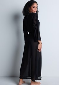 Robe longue noire transparente avec des manches longues, une élégante fluidité et une taille nouée. Texture lisse, design jusqu'aux chevilles et esthétique minimaliste.