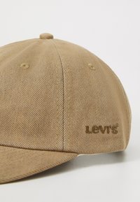 Homokszínű baseball sapka texturált anyagból, ívelt karimával, varrott díszítésekkel és barna hímzett "Levi's" logóval.