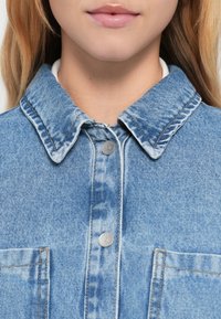 Denimskjorte i falmet lyseblå farge, stor krage, to frontlommer og sølvfargede trykknapper. Teksturen virker lett slitt.