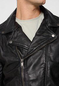 Veste en cuir noir de biker avec un col à revers, une fermeture éclair asymétrique et des boutons-pression métalliques. Texture lisse et design ajusté.