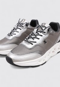 Chaussures de sport argentées avec une tige en mesh, une forme arrondie, des détails texturés, des lacets noirs et une semelle blanche à motifs.