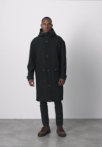 Czarny długi płaszcz z kapturem, z przodu guziki, liczne kieszenie i luźny krój. Noszony na ciemnych spodniach i brązowych butach.