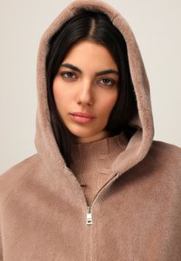 Lysebrun zip-up hættejakke med en luksuriøs tekstur, der har en stor hætte og et kontrastfarvet strikket indre lag i en dybere brun nuance.