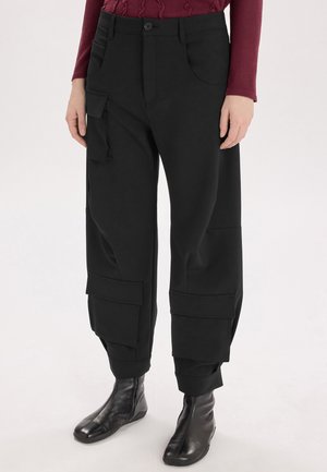 Persona che indossa pantaloni cargo neri con grandi tasche e stivaletti neri, mostrata dalla metà del busto fino ai piedi su sfondo bianco.