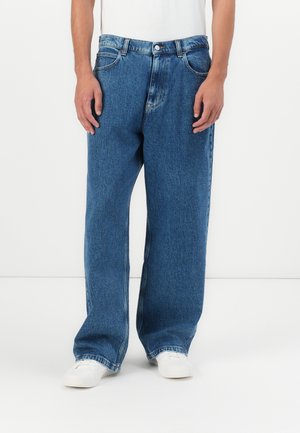 Uomo che indossa jeans larghi di denim blu e sneakers bianche, in piedi contro uno sfondo bianco uniforme.