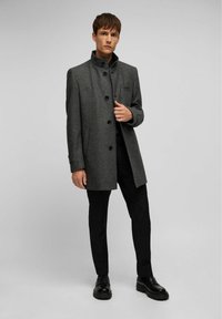 Grijs visgraatjas met een hoge kraag, knoopsluiting en zijzakken. Gecombineerd met zwarte broek en stevige zwarte schoenen.