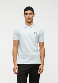 Jovem em pé, vestindo uma camisa polo azul-clara com um pequeno logotipo preto e branco no peito e calças pretas.