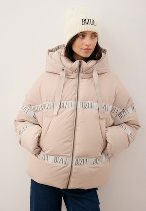 Gewatteerd jack in lichtbeige met capuchon, voorzien van witte bandjes met daarop "BIZUU" gedrukt en een voorsluiting met ritssluiting. Gecombineerd met een witte geribbelde muts.