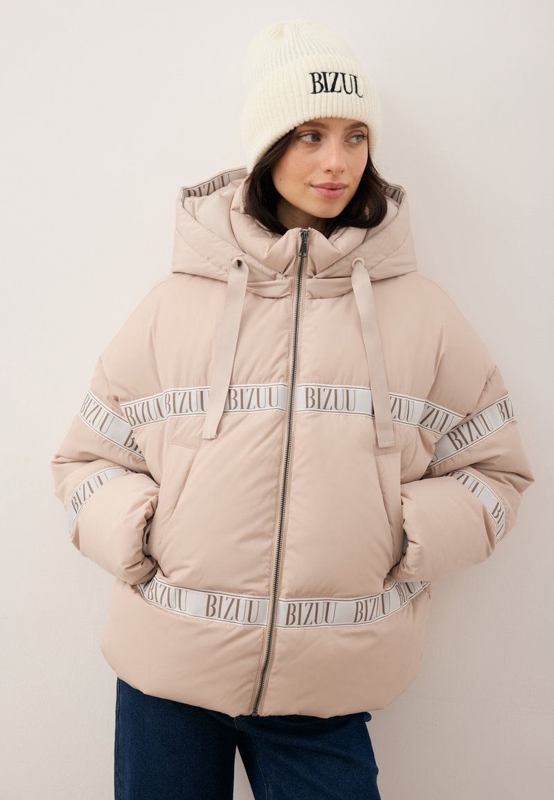 Kurtka puchowa w jasnym beżu z kapturem, ozdobiona białymi paskami z nadrukiem "BIZUU" oraz przednim suwakiem. Do tego biały prążkowany beanie.