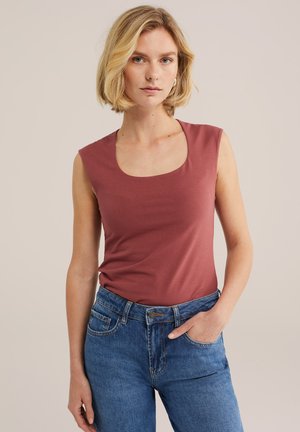 Donna bionda che indossa un top senza maniche color rosa polvere e jeans blu a vita alta, in piedi con una mano nella tasca davanti a uno sfondo semplice.