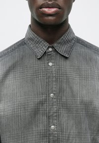 Grå rutig skjorta av denimtyg med knäppkrage, utrustad med silverknappar och ett texturerat mönster.